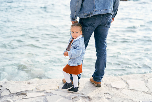 Dad in a denim jacket and jeans holds a little girl by the hand, standing on the rocks by the water. Girl turned to face the camera - Фото, изображение