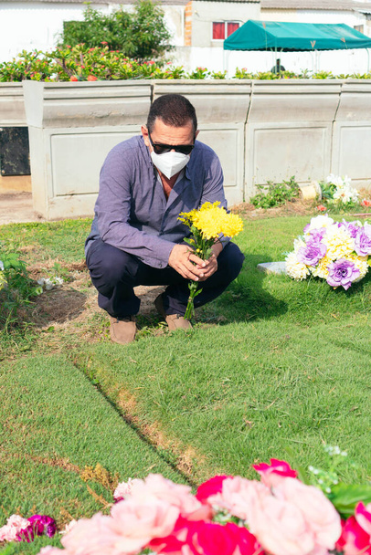 Μεγαλύτερος άντρας με προστατευτική μάσκα που τοποθετεί λουλούδια σε τάφο. Επιδημία του κορωναϊού. - Φωτογραφία, εικόνα