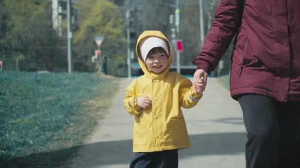 Sarı yağmurluklu çocuk şehirde yürüyor ve annesinin elini tutuyor. - Video, Çekim