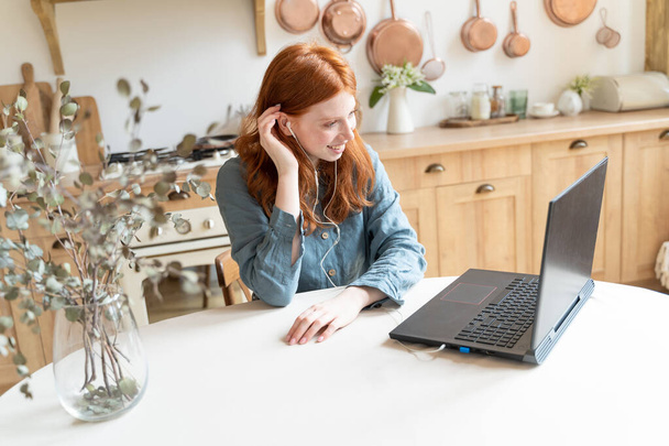 Happy red haired woman Online video call using laptop at kitchen. Red haired white woman having online conference smiling enjoying conversation. Long distance relationship. Stay home freelance remote  - Foto, afbeelding