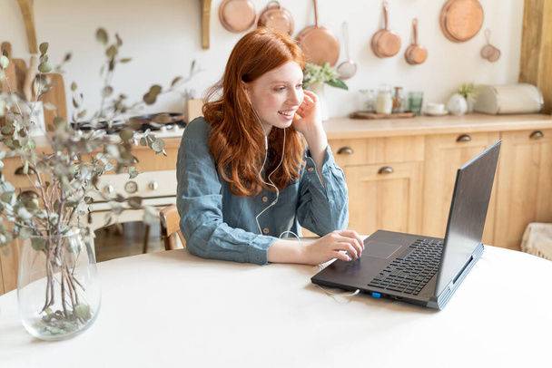 young red haired Freelancer girl using laptop and headphones having online conference chatting video call at home kitchen. Working remotely from home. Distance communication blogger conference - Foto, imagen
