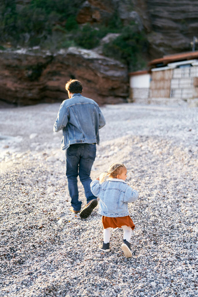 La bambina corre dietro a papà sulla spiaggia di ciottoli sullo sfondo di montagne rocciose. Vista posteriore - Foto, immagini