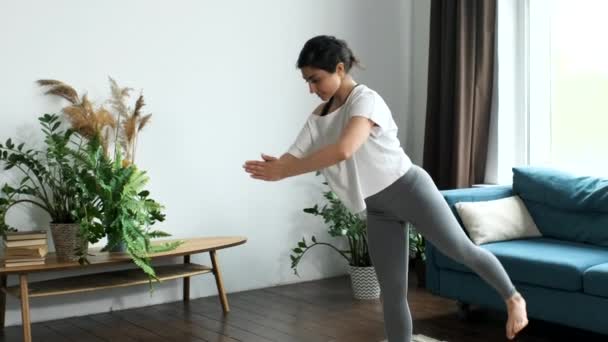 Genç atletik kız evde tek ayak üstünde durarak yoga dersleri alıyor. Evde, parlak bir odada, vücut esneme hareketleri yapıyor. - Video, Çekim