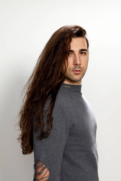 Привлекательный молодой парень с очень длинными вьющимися волосами позирует в студии. Стиль, тенденции, концепция моды. - Фото, изображение