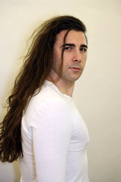 Ελκυστικός νεαρός με μακριά σγουρά μαλλιά ποζάρει στο στούντιο. Στυλ, τάσεις, έννοια της μόδας. - Φωτογραφία, εικόνα