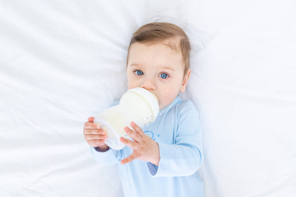 baby boy eats milk from a bottle on the bed before going to bed in a blue bodysuit, baby food concept - Foto, afbeelding