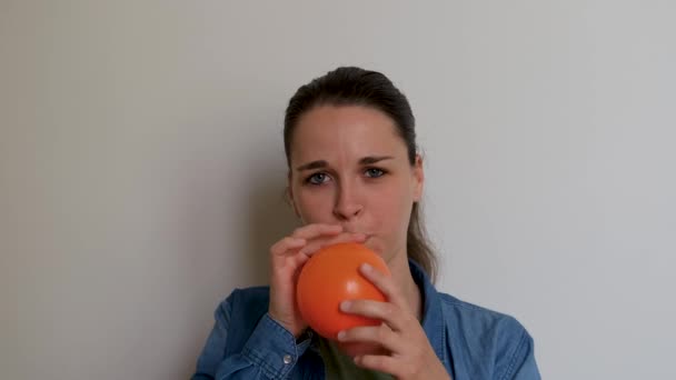 Jonge mooie vrouw van Kaukasische Europese uiterlijk blaast oranje ballon met haar mond op witte achtergrond. Meisje bereidt zich voor op vakantie en blaast ballon op en laat het dan wegvliegen. - Video