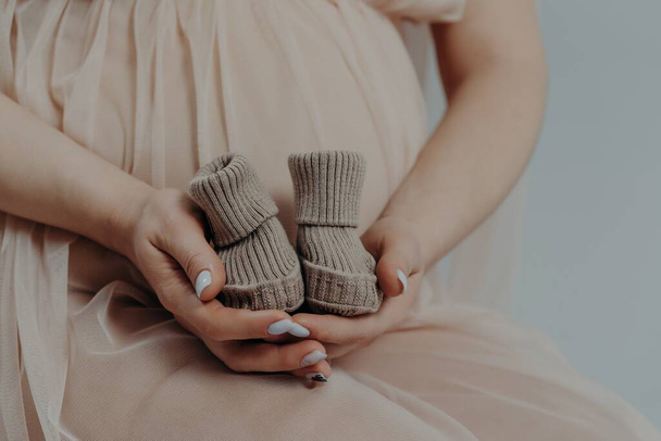 Koncepcja macierzyństwa i ciąży. Oczekująca matka przygotowuje się do porodu dziecka trzyma buty w rękach stwarza kryty jest na trzecim trymestrze ciąży - Zdjęcie, obraz