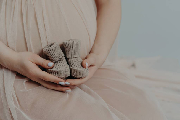 Concept de maternité et de grossesse. Femme enceinte méconnaissable tient de petites bottes sur le ventre porte une robe de fête. Sans visage future mère - Photo, image