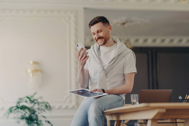 Щасливий чоловік фрілансер дивиться на телефон і посміхається, сидячи на робочому столі
 - Фото, зображення