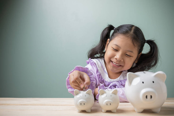 Egy ázsiai kislány érmét tett egy fehér malacperselybe otthon, egy fa asztalon. Takarítson meg pénzt tandíjra, az iskolai felszerelések költségére a jövőben. Ötletek a jövőbeli oktatási célú megtakarításra. - Fotó, kép