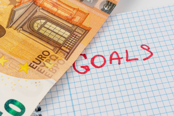 Word GOALS is written in red on a checkered paper notepad. Fifty euro banknote opens an inscription close up. Wealth goal definition and achievement concept. Financial prosperity dream idea - Fotoğraf, Görsel