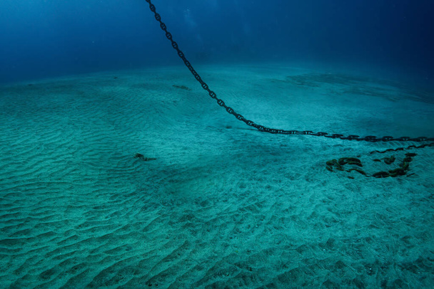 Foto submarina de una pesada cadena de anclaje de acero que cuelga de la superficie del agua y yace en el fondo de arena blanca, aguas azules turbias más profundas en el mar tropical. - Foto, imagen