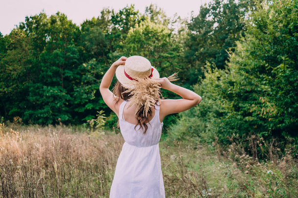 Девушка в соломенной шляпе спиной к фотографу. Девушка в белом платье в летний солнечный день. - Фото, изображение