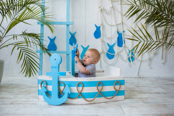 bebé niño rubia en un traje a rayas se sienta en un barco azul y blanco sobre un fondo con una decoración de mar. Viajes de verano con un niño - Foto, imagen