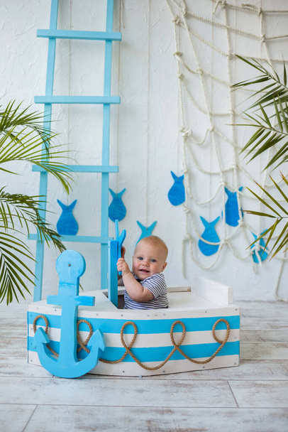 縞模様のスーツの白人の赤ん坊は海の装飾が付いている背景の青そして白のボートに坐る。垂直方向 - 写真・画像
