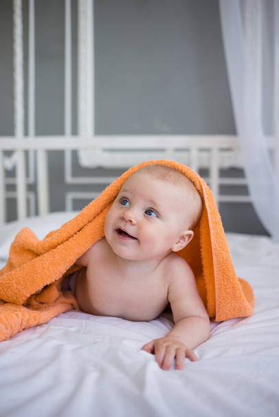 naakt jongetje ligt onder een oranje handdoek op het bed. Babyverzorging - Foto, afbeelding