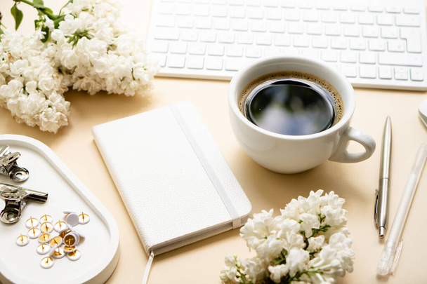 Beige and white work space with coffee cup, notepad and lilac flowers - Foto, Bild