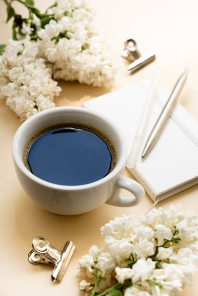 Μπεζ και λευκό χώρο εργασίας με φλιτζάνι καφέ, σημειωματάριο και πασχαλινά λουλούδια - Φωτογραφία, εικόνα