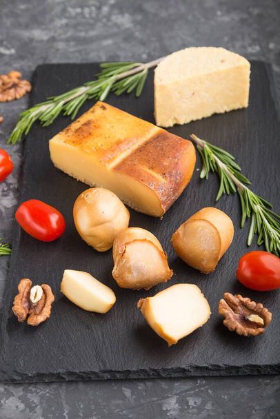 Smoked cheese and various types of cheese with rosemary and tomatoes on black slate board on a black concrete background. Side view, close up, selective focus. - Photo, image