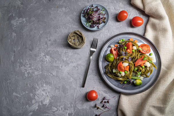 Ταλιατέλ πράσινο σπανάκι ζυμαρικά με ντομάτα, μπιζέλι και λαχανάκια σε γκρι φόντο μπετόν και λινό ύφασμα. Κάτοψη, επίπεδη lay, αντίγραφο χώρου, σκληρό φως. - Φωτογραφία, εικόνα