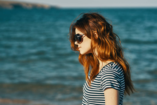 roodharige vrouw met zonnebril op het strand bij de blauwe zee en bergen op de achtergrond - Foto, afbeelding