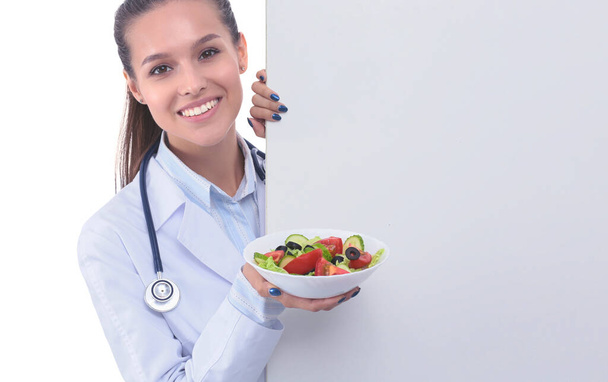 Portrait of a beautiful woman doctor holding a plate with fresh vegetables standing near blank. Woman doctors - Photo, Image