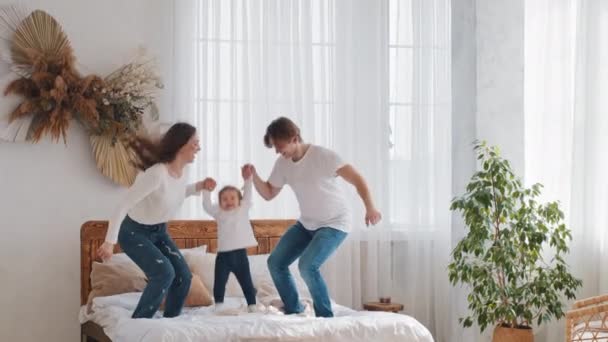 Young parents married couple caucasian family with little daughter toddler child girl jumping on bed holding hands jump into air in bedroom having fun playing at home enjoy parenting and communication - Footage, Video