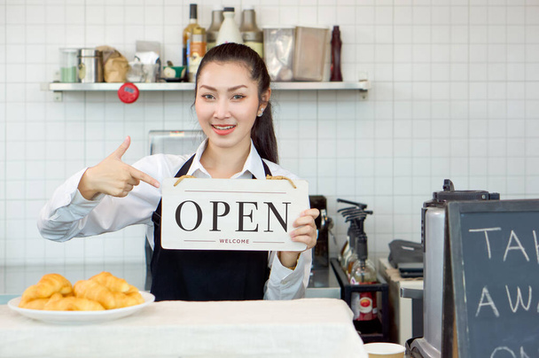Jonge Aziatische winkelier met een glimlach houdt een OPEN bord voor een coffeeshop teller. Ochtendsfeer in een coffeeshop. - Foto, afbeelding