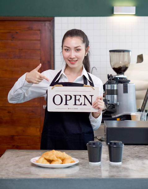 Giovane negoziante asiatico con un sorriso tiene un cartello OPEN davanti a un bancone di una caffetteria. Atmosfera mattutina in una caffetteria. - Foto, immagini
