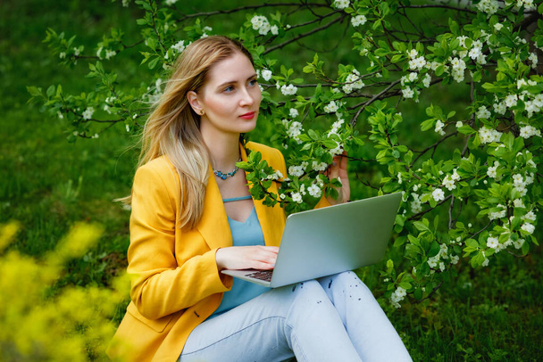 Sarı ceketli güzel bir kadın çimlerin üzerinde oturur ve çiçeklenme mevsiminde parkta dizüstü bilgisayarıyla çalışır. Serbest çalisma, uzaklik ögrenme veya uzaktan çalisma konsepti. - Fotoğraf, Görsel