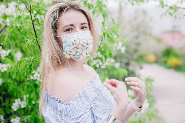 A girl in a medical mask decorated with live flowers poses in flowering trees - Foto, Imagem