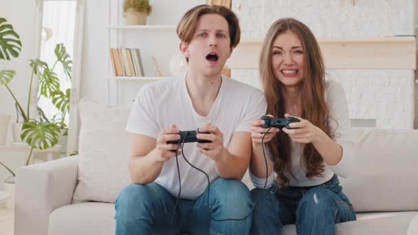 Παντρεμένο ζευγάρι καυκάσιος γυναίκα και χιλιετή άντρας φίλος και φίλη παίζει κονσόλα online ανταγωνισμός αγώνα βιντεοπαιχνίδι κάθεται στον καναπέ στο σπίτι κορίτσι χάνει αναστατωμένος δείχνει γλώσσα στον νικητή άντρα - Πλάνα, βίντεο