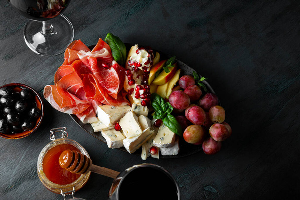  A set of appetizers for wine, jamon, pepperoni, cheese, grapes, peach and olives on a plate top view. Snack board and two glasses of red wine on dark blue background, free space for text - Fotoğraf, Görsel