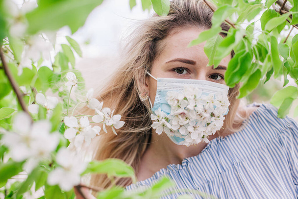 A girl in a medical mask decorated with live flowers poses in flowering trees - Фото, изображение