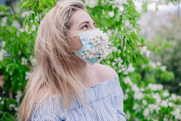 A girl in a medical mask decorated with live flowers poses in flowering trees - Foto, afbeelding