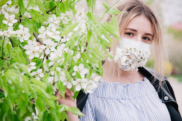 Ένα κορίτσι με ιατρική μάσκα διακοσμημένο με ζωντανά λουλούδια ποζάρει σε ανθισμένα δέντρα. - Φωτογραφία, εικόνα