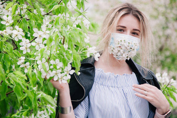 A girl in a medical mask decorated with live flowers poses in flowering trees - Φωτογραφία, εικόνα