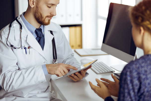 Bearded doctor and patient woman discussing current health examination while sitting and using tablet computer in sunny clinic, close-up. Medicine concept - Photo, image