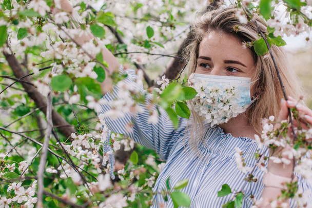 A girl in a medical mask decorated with live flowers poses in flowering trees - Fotoğraf, Görsel