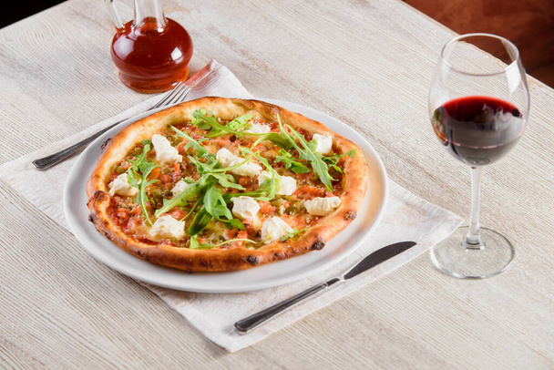 Pizza lazaccal, feta sajttal, szósszal, paradicsommal, fehér kerek tányéron, világos fa asztalon, üveg vörösbor hátterén - Fotó, kép