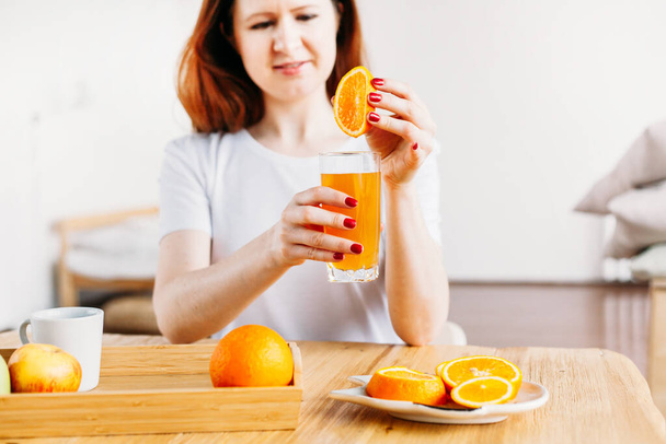 Avrupalı görünümlü bir kız masada beyaz bir tişörtle oturuyor, meyve suyu ve meyve suyu yapmak için portakallar, vitaminlerin faydaları, sabah - Fotoğraf, Görsel