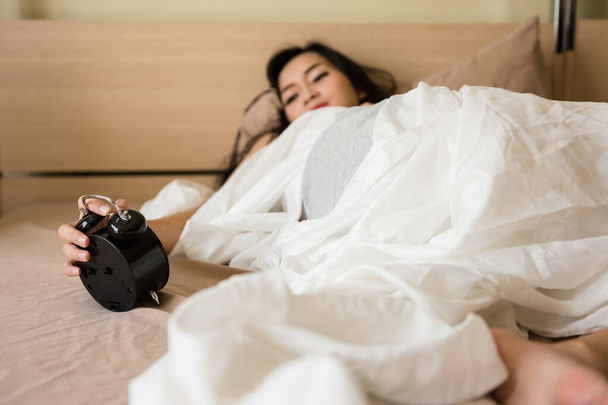 Asyalı bir kadın, 20-30 yaşlarında, sabah yatarken saati durdurmuş. Uyanma, banyo yapma, giyinme ve işe gitme zamanı.. - Fotoğraf, Görsel