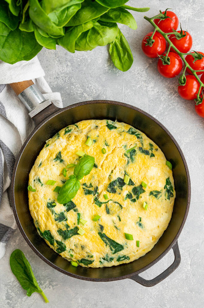 Omelet met spinazie, groene uien en feta kaas in een gietijzeren koekenpan op een grijze betonnen ondergrond. Gezond ontbijt. Bovenaanzicht, kopieer ruimte - Foto, afbeelding