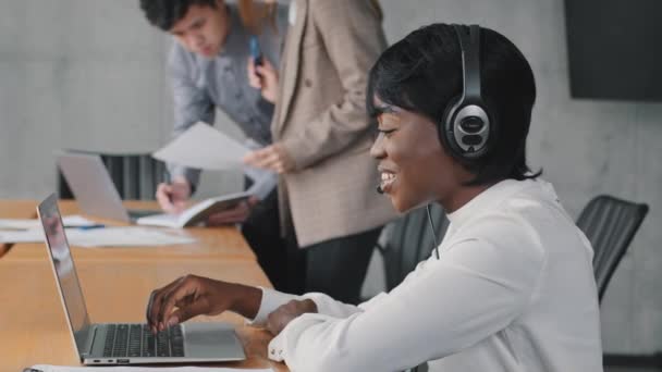 Sonriente africano agente de centro de llamadas femenino usar auriculares inalámbricos usando portátil mirando a cliente de consulta de pantalla con problema en línea hacer videollamada en la oficina, negro mujer operador servicio de apoyo - Imágenes, Vídeo