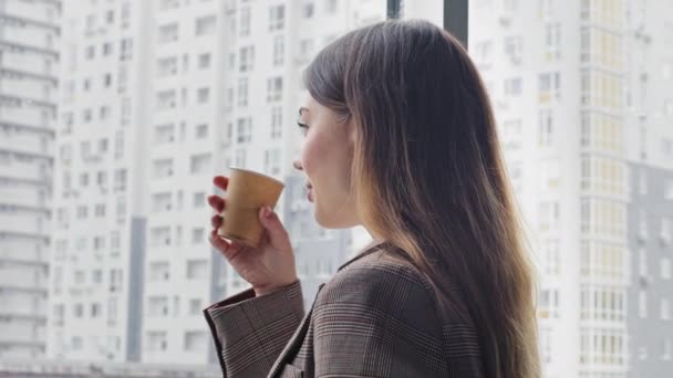 Kaukasische jonge duizendjarige zakelijke vrouw leider baas specialist professionele stands in kantoor kijken uit raam op wolkenkrabber stad drinkt thee koffie uit kartonnen beker verandert hoofd kijkt in camera - Video