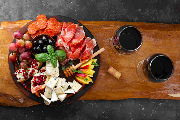  Un conjunto de aperitivos para el vino, jamón, pepperoni, queso, uvas, melocotón y aceitunas en una tabla de madera vista superior. Snack board y dos copas de vino tinto sobre fondo gris oscuro - Foto, imagen