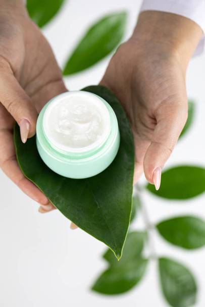 Close-up frasco de creme em folha verde fresca em mãos sobre fundo de ervas brancas - Foto, Imagem