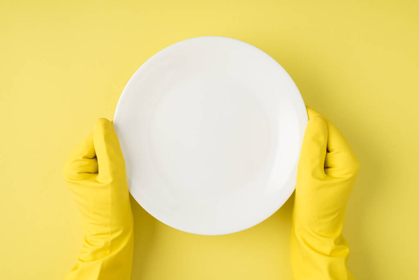 Top view photo of hands in yellow rubber gloves holding white clean plate on isolated yellow background with copyspace - Fotó, kép