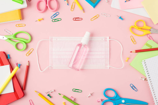 Фотография многоцветных канцелярских ножниц ручки карандаши кнопки зажимы калькулятор копировальные книги и дезинфицирующая бутылка на медицинской лицевой маске в середине изолированных пастельно-розовый фон - Фото, изображение